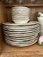 Stack of ceramic bowls and dinner plates with pink rose pattern on wooden shelf.