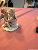 Front and side view of the porcelain figurine with two cherubic angels seated and embracing on a textured base.