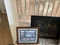 Framed mountain print leaning against a brick fireplace on a tiled hearth with dog bowls nearby.