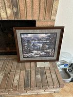 Framed mountain landscape print shown leaning against a brick fireplace, displaying a detailed scene of a snowy mountain range, pine trees, and a body of water under a cloudy sky. Frame is brown wood with a gray textured mat surrounding the print.