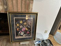 Framed print of a floral still life with colorful flowers and black-gold frame leaning against a brick fireplace.