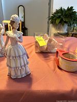 Photo showing porcelain lady figurine in white dress with purple ruffles, along with packaging materials and tape in the background.
