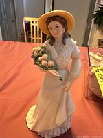 Front and side view of the porcelain figurine of a woman with a wide-brimmed hat holding pink flowers.