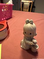 Portrait of the Precious Moments figurine showing the girl with a bow in her hair holding a cup and teapot on a table