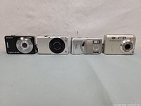 Four digital cameras in a row showing front sides: Sony Cyber-shot, one unknown white and silver camera, Vivitar ViviCam 5110, HP Photosmart M537