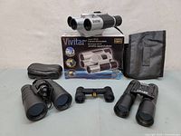 Four pairs of binoculars displayed on neutral background including Vivitar Digital 10x25 camera binocular with box and soft case