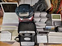 Photo showing assortment of electronics on table; includes white MacBook, two portable DVD players, weather clock, keyboards, jewel cases, boombox, and remote