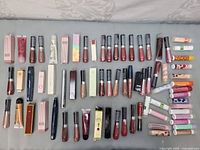 Overview photo showing an assortment of lip glosses, lip conditioners, perfumes, and lip balms arranged in rows on a fabric surface