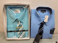 Two men's XL dress shirts in new condition. Left is Alessio brand light blue pleated front shirt in packaging with striped tie and tie clip. Right is INC brand solid light blue shirt with striped tie and pocket square, tagged.