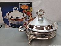 Vintage silver-plated casserole dish with ornate handles and feet, a lid with decorative floral handle, and Pyrex glass insert visible next to the box.