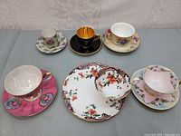 Full view of six vintage teacups with saucers showing diverse floral and gold-accented designs on a gray surface.