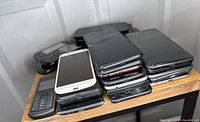 Stack of various smartphones mostly black, visibly cracked screens, older and newer models.