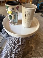 Two Starbucks glass cups on a white tray. One is white with black Starbucks logos and checkboxes, the other colorful with illustrations, missing top lid.