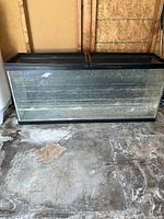 Front view of the large aquarium tank showing glass panels, black framing, and sliding lids on top, placed on cement floor against a plywood wall.