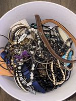 Photo shows a bucket filled with a tangled assortment of women's jewelry including metal bracelets, pearl and bead necklaces, leather strap, and other varied costume jewelry pieces.