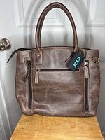 Front view of brown genuine leather purse displaying two vertical zippered compartments, sturdy handles, and metal details on sides.
