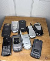 Photo showing 8 vintage flip phones arranged on wooden surface revealing various brands and styles including flip, candy bar with visible antennas and camera