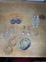 Top down photo showing six pairs of earrings arranged on wooden surface, featuring multiple styles including hoops, dangling, and custom yarn wrapped hoops.