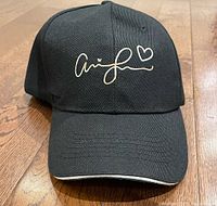 Front view of black baseball cap with white stylized signature and heart printed on front panel. Shows curved brim with white edge detail.