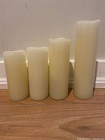 Four battery operated candles standing upright against a wall on a wood floor, showing different heights and off-white color.
