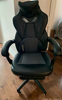 Front view of black gaming chair showing padded arms, mesh lumbar cushion, headrest, and leather-like upholstery.