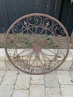 Front view of a round decorative metal garden wheel with intricate scroll and floral designs and rust brown finish against outdoor garden background.