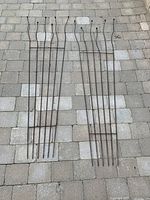 Two brown metal garden trellises laid flat on a concrete paver surface showing vertical bars, horizontal connectors, and decorative rounded finials on top.