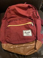 Front view of burgundy Herschel knapsack showing leather bottom and front zipper pocket with yellow trim.