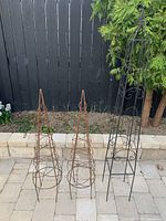 Two rust-colored conical iron trellises placed on patio against dark fence and garden bed behind