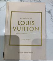 Image shows white hardcover book with gold embossed title 'Little Book of Louis Vuitton' by Karen Homer, subtitle 'The story of the iconic French house', marked as unofficial and unauthorized.