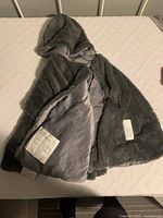 Grey soft massage cape with hood laid open on a bed, showing plush exterior and smooth interior lining. Battery pack placed on cape.