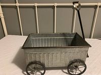 Small rectangular galvanized metal wagon placed on a white surface with a beige metal headboard background. The photo shows the side view, highlighting the metal handle and four metal fixed wheels.