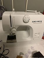 Front view of white Euro-Pro mini sewing machine showing stitch selector, thread guides, needle area, and control buttons.
