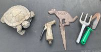 Top-down view of the turtle figurine, metal dog sign, hose faucet, and two garden hand tools on a dark surface.