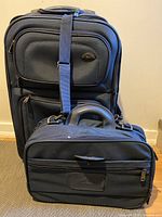Photo showing the upright mid-size suitcase behind the carry-on travel bag, both in dark canvas with wear visible.