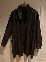 Front view of dark chocolate brown leather jacket hung on a wooden hanger showing button closure and deep pocket details.