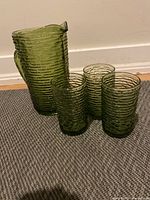 Photo showing the green ribbed glass pitcher with three matching small glasses on a carpeted floor showing size and condition.