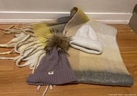 Wide shot showing the giant multicolor scarf, the white tuque, and the purple cashmere hat with fur pom pom all spread out on the floor.