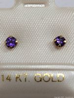 Close-up photo showing pair of small round amethyst earrings set in 14KT gold prong settings in white jewelry display.
