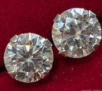 Close-up photo of two faceted round moissanite stones in 10KT gold prong settings on red background.