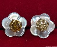 Close-up image of earrings showing flower shape with white mother of pearl petals and yellowish diamond center, on red background.
