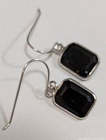 Close-up image of both sapphire earrings with silver hooks displayed on a white surface, showing faceted deep dark blue rectangular stones set in white gold.
