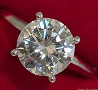 Close-up front view of the 14KT white gold ring with large round moissanite gemstone mounted in four prongs against a red background.