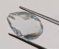 Close-up front view of light blue aquamarine gemstone held with tweezers showing pear shape and faceting.