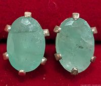 Close-up photo of two oval emerald earrings set in silver prong mounts with a red background.
