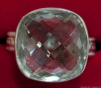 Close-up photo of silver ring with cushion-cut green amethyst gemstone showing facets and reflection on red background.