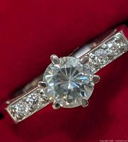 Close-up photo of the silver ring showing the round 1.65ct moissanite center stone and three smaller stones on one side of the band, against a red background.