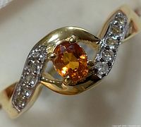 Close-up photo of silver ring with central orange sapphire and clear stones on band, showing prong setting and twisted band design.