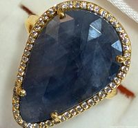 Close-up of pear-shaped bluish sapphire gemstone with diamond halo on gold-tone band