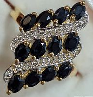Close-up front view showing oval faceted blue sapphire stones and two rows of white topaz stones in curved lines on silver ring with gold-tone prongs and band.